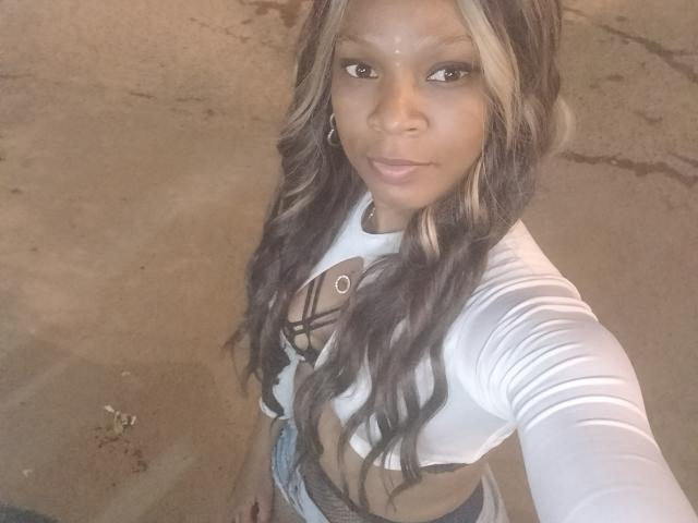 Haddie with long, wavy blonde and brown hair, wearing a white, cut-out top showing her tits, and blue jeans, taking a selfie, looking directly at the camera.