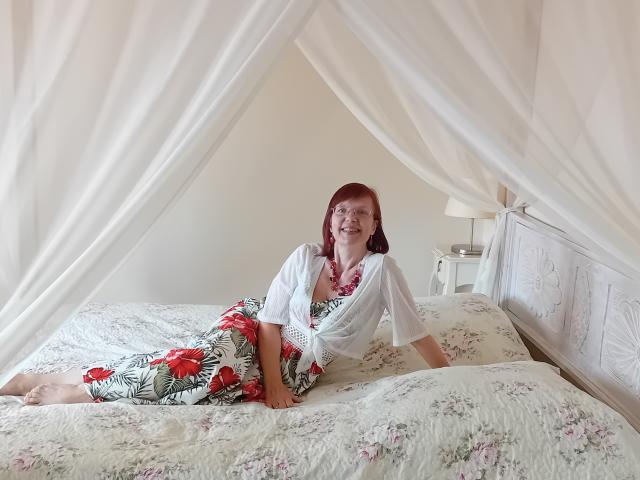 JuicyLady69 with red hair, wearing floral pants and a white shirt, lying on a floral bed, smiling, white sheer canopy, white bed, white nightstand, white lamp.