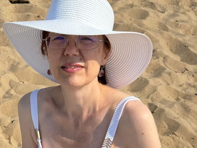 JuicyLady69 with white sunhat, pink glasses, and white bikini top, showing light skin and small tits, smiling at the camera on a sandy beach.