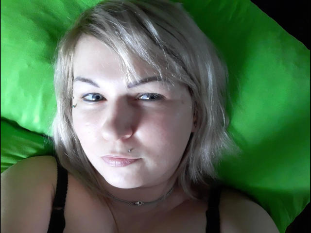 LiebeMauschen-hot with short blonde hair, nose piercing, and black bra, lying on green pillow, looking at camera with slight smile. Her tits are visible, choker around her neck.