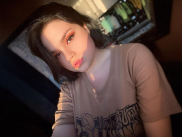 AdelinaOlsen with pale skin, pink lipstick, and brown eyes, wearing a gray "Riot" t-shirt, sunlight on her face, short brown hair, slightly tilted head, soft focus.