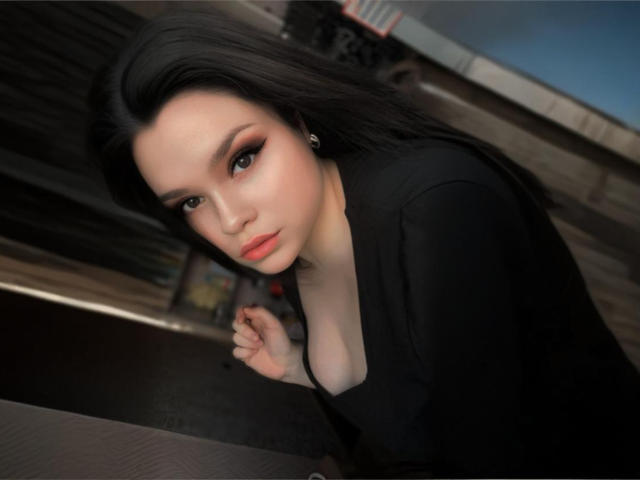 AdelinaOlsen with pale skin, black hair, and orange lipstick, leaning forward in a black top, showing a small amount of her tits, with a serious expression.