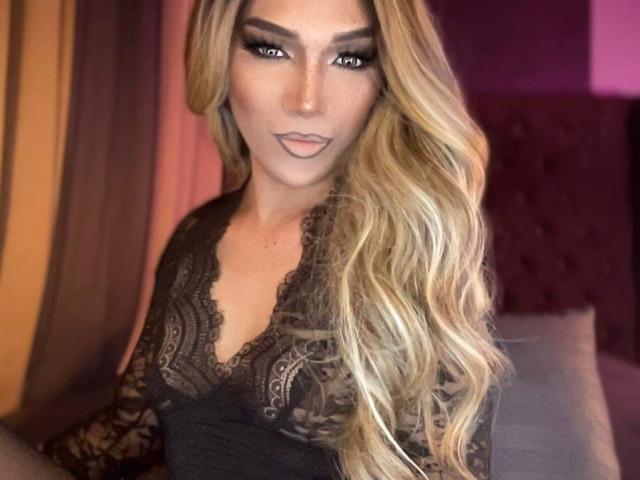 Lola69Xx, con cabello rubio largo, una blusa de encaje negro y maquillaje de ojos ahumados, mostrando sus labios carnosos y sus pechos en una luz suave y cálida..