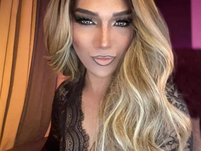 Lola69Xx ha lunghi capelli biondi ondulati, eyeliner scuro e rossetto nude, indossa una maglia nera in pizzo, con un'espressione seria e le labbra leggermente dischiuse. I suoi occhi sono intensi e penetranti..