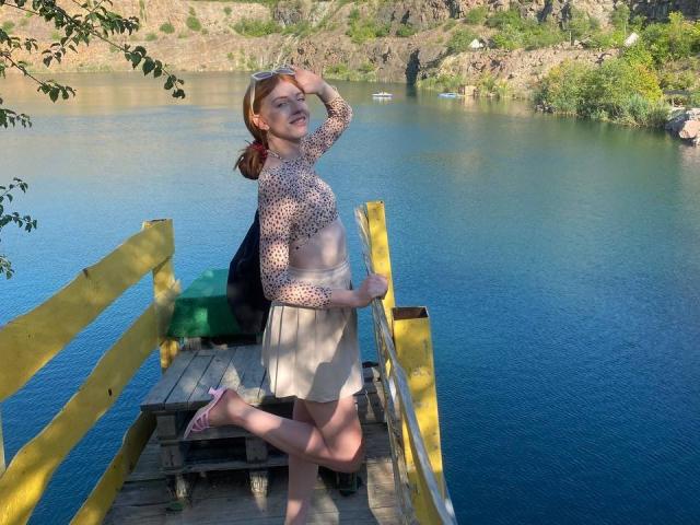 Eine GingerFairy mit roten Haaren, die ein Top mit Punkten und einen beigen Rock trägt, steht auf einem gelben Holzsteg, lächelt und hat ihre Hand auf dem Kopf, wobei ihr Hintern zu sehen ist..