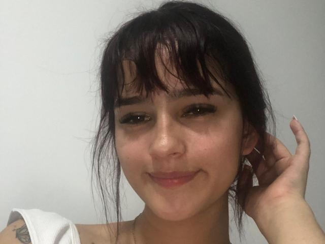 MelodyMansson has dark brown hair with bangs, a cute nose ring, and a subtle smile. She's touching her ear with one hand, showing a tattoo on her shoulder.