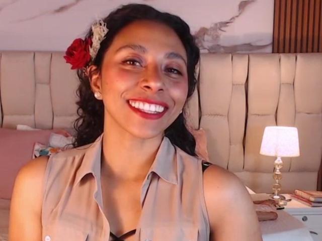 JanEvans has dark curly hair with a red flower, wearing a beige sleeveless blouse, smiling with red lipstick, and a warm light in the background.