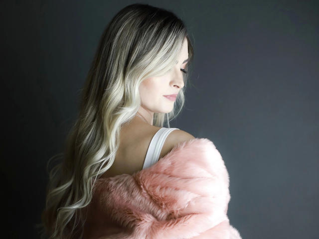 LanaFire – atemberaubend mit langen, welligen blonden Haaren, einem weißen Tanktop und einem pinkfarbenen, fellbesetzten Mantel, der von der Schulter fällt, blickt sie nach unten mit zartem, pinkfarbenem Lippenstift und einem ruhigen, verführerischen Ausdruck..