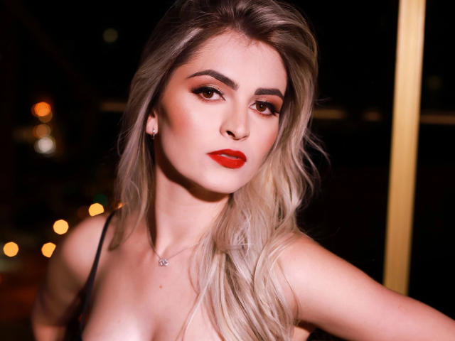 LanaFire-hot a les cheveux blonds, du rouge à lèvres, et regarde l'appareil photo avec un léger sourire, montrant ses seins dans un haut noir à bretelles, la nuit..