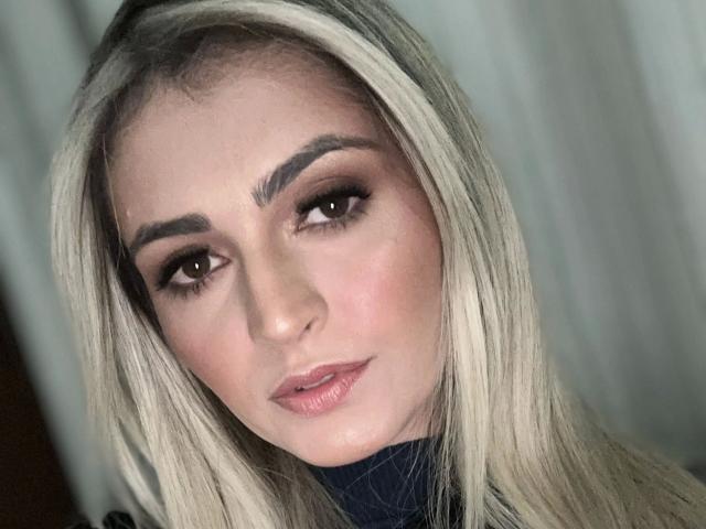 LanaFire hat blondes Haar, dunkle Augenbrauen, braune Augen und rosafarbene Lippen. Ihr Gesicht ist leicht geneigt, und sie blickt mit einem subtilen, verführerischen Ausdruck in die Kamera..