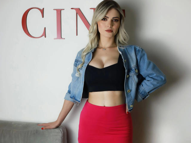 Lana, mit blonden Haaren, schwarzer BH, der ihre Brüste zeigt, einer blauen Jeansjacke und einem roten Rock, steht mit einer Hand auf einem grauen Sofa und blickt in die Kamera..