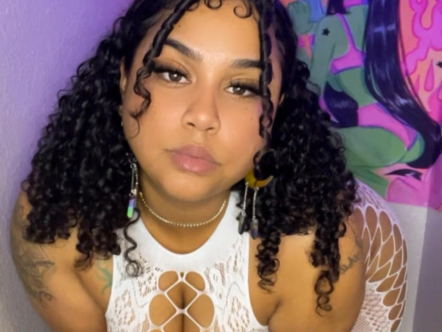 KushandRoses has curly black hair, brown eyes, and is pouting with pink lips. She's wearing a white lace top, showing her tits, with tattoos on her arms.