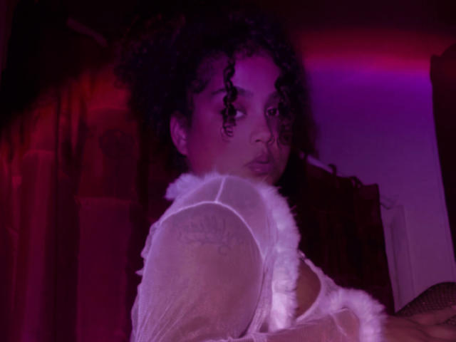 KushandRoses with curly black hair, wearing a white sheer top with fluffy trim, purple and red neon lighting, looking over shoulder, medium shot. Boobs partially visible.
