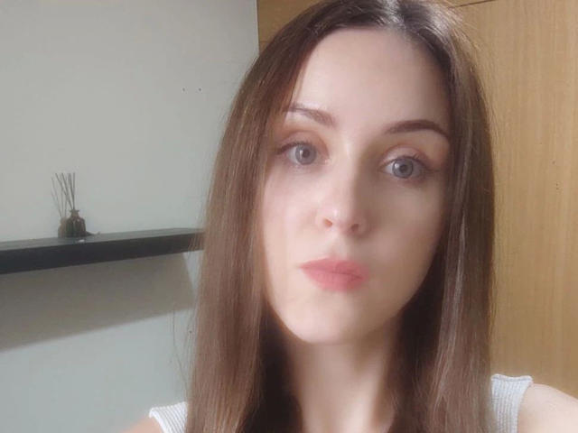 KnackigeOssi-hot has pale skin, straight brown hair, blue eyes, and pink lips, wearing a white top, looking at the camera with a neutral expression.