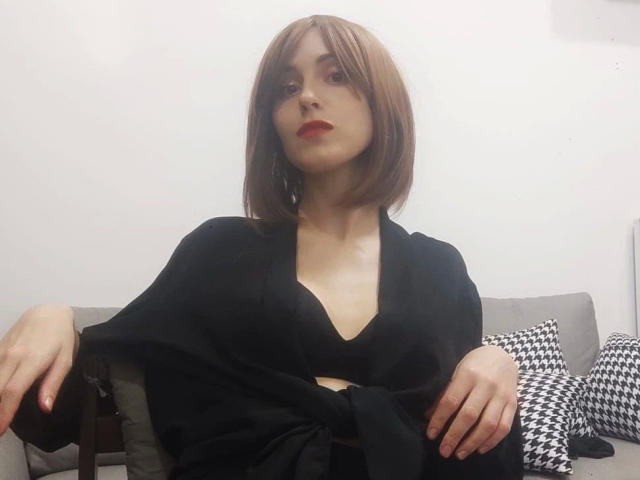 KnackigeOssi-hot with a bob haircut, red lipstick, black top, and black shawl, sitting on a gray sofa with black-and-white pillows, hands resting on her knees.