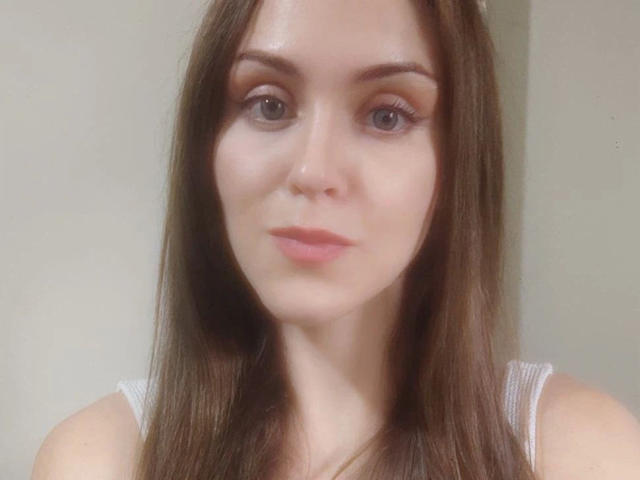 KnackigeOssi-hot has long brown hair, light skin, and small tits. She's looking at the camera with a subtle smile, wearing a white tank top.