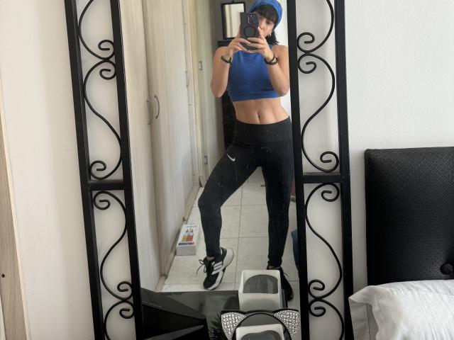 SeleneBrigth in a mirror selfie, wearing a blue crop top, black tight pants, black and white sneakers, and a blue cap, showing her toned abs and slim waist.