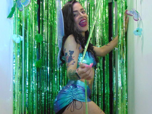 LunaRoose with long dark hair, blue-green shiny dress, tattoos, and pink nails, sticking tongue out, holding green streamer, standing in front of green metallic curtain.