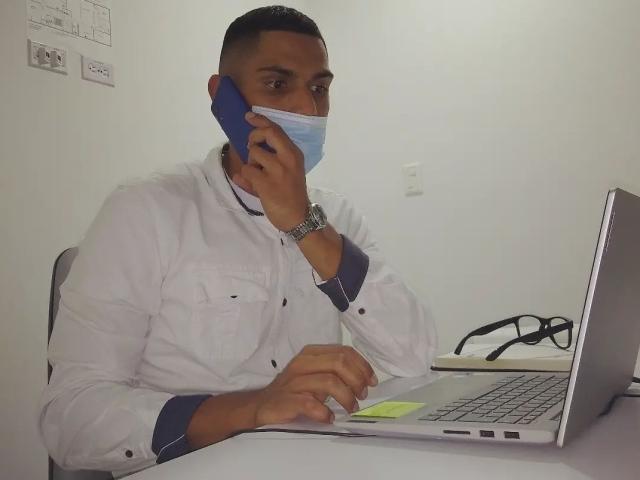 McBride with a blue mask, white lab coat, and watch, holding a phone to his ear, typing on a laptop with yellow sticky notes, glasses on the right.