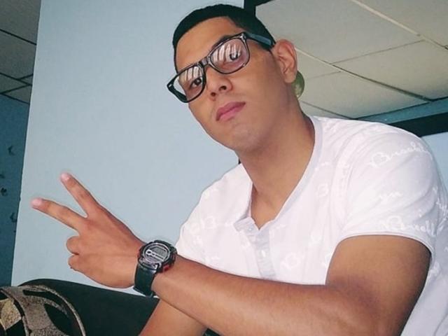 McBride, a short-haired dude with glasses, wearing a white shirt and black wristwatch, makes a peace sign with his right hand, looking slightly annoyed.