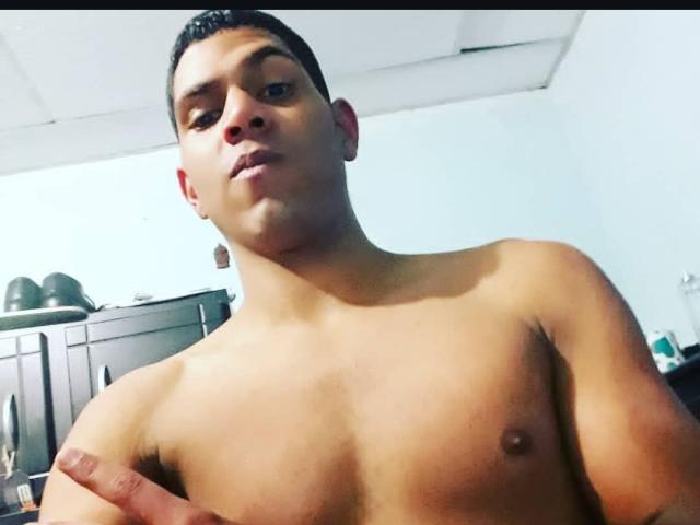 McBride, shirtless, with short dark hair, looking at camera, medium brown skin, small nipples, left ear earring, right hand on chest, in dimly lit room.