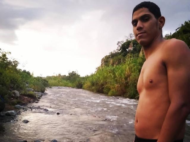 McBride, shirtless, standing by a flowing river, has a muscular, tan, flat-chested torso, short dark hair, and a serious expression, in a nature setting with greenery.