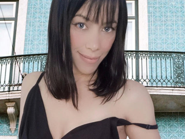 Chloe Vieira, aantrekkelijk met zwart, halflang haar, een pony en blauwe ogen, draagt een zwarte top met een open schouder, waardoor haar kleine borsten zichtbaar zijn, en ze glimlacht zachtjes..