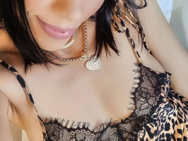 El rostro de ChloeVieira-hot, con lápiz labial rosa, cabello negro hasta los hombros, vistiendo un top de encaje negro con estampado de leopardo y un collar con forma de corazón y una cadena. Se ven los senos..