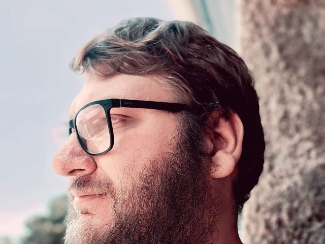 LucienFrenchy with a beard, black rectangular glasses, and wavy brown hair, standing near a tree, eyes closed, looking content. His nose is prominent and his expression is calm.