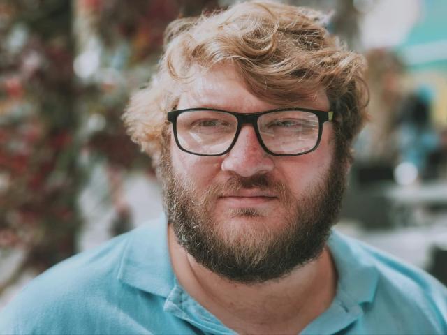 LucienFrenchy has a curly blonde beard, black glasses, and a slightly chubby face with a blue shirt, looking slightly annoyed. His nose is red and he's outdoors.