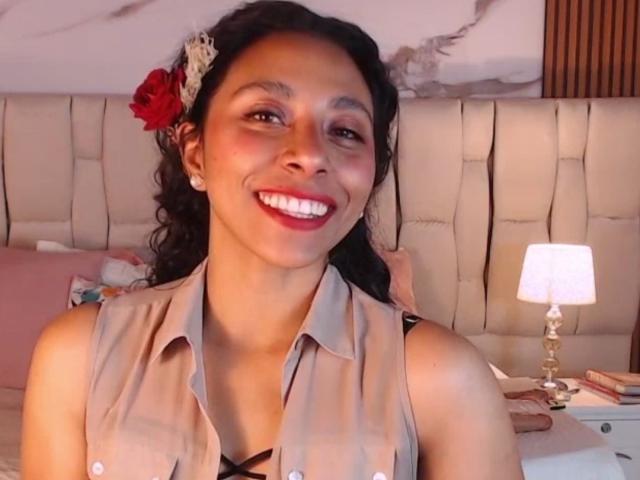 JanEvans has dark curly hair with a red flower, wearing a beige sleeveless shirt, smiling with red lipstick, and showing white teeth. Her ass is partially visible.