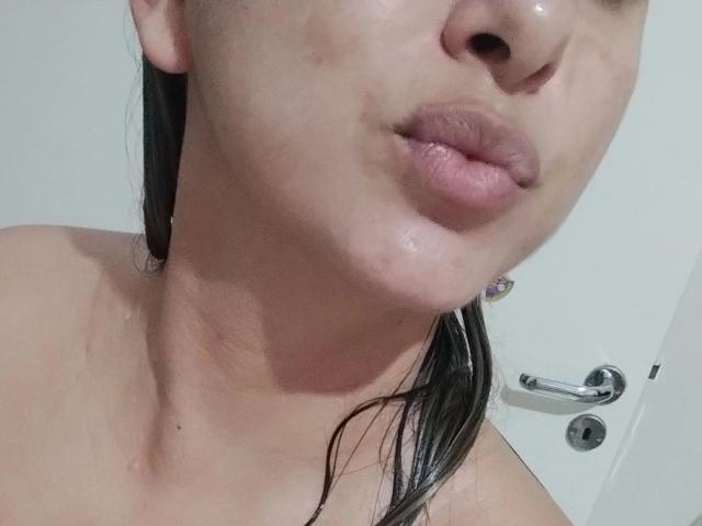 KatePink-hot's face with wet skin, slightly parted lips, and black wet hair. Her pinkish lips have a shiny, wet look. Her collarbone is visible, and a door is behind.