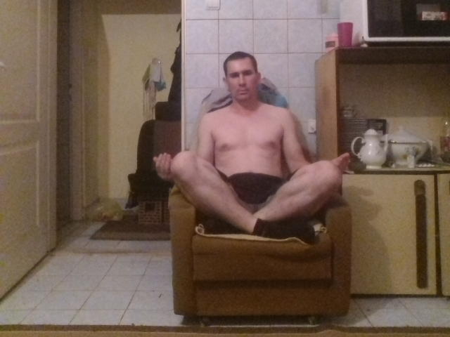 Zelkic, ohne Hemd, sitzt in einem braunen Sessel, die Beine gespreizt, die Hände auf den Knien, in einer Küche, mit ernstem Gesichtsausdruck, trägt schwarze Shorts, weiße Socken und ein Handtuch auf dem Kopf..