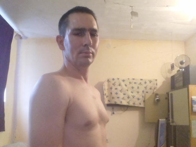 Zelkic, senza camicia, con un'espressione seria, piccoli seni, capelli scuri e corti, pelle chiara, in piedi in una stanza, che guarda la telecamera, leggermente muscoloso, con una luce sul soffitto visibile..