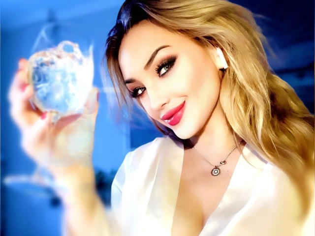 Ohyeahhh, linda com cabelo loiro, segurando uma esfera de vidro transparente, vestindo uma camisa branca, sorrindo, com batom rosa, usando um colar, fundo azul, olhando para a esfera com uma expressão divertida..