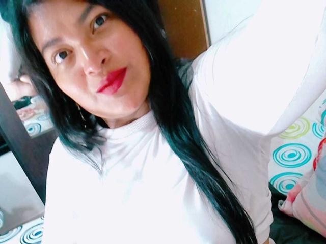 KamilaSquirt has long black hair, red lipstick, and a white shirt, with a slightly flushed face in a selfie. Boobs are not visible. She's smiling at the camera.