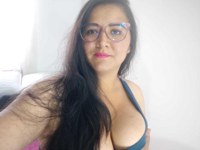 Philipa with long black hair, wearing glasses and a dark blue halter top, showing her big tits, pink lipstick, and a slight smile, in a bright room.