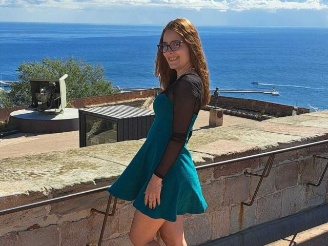 LucyLucia with red hair, glasses, and a teal dress with black sleeves, standing on a stone balcony, looking over her shoulder, showing her ass. Ocean in the background.