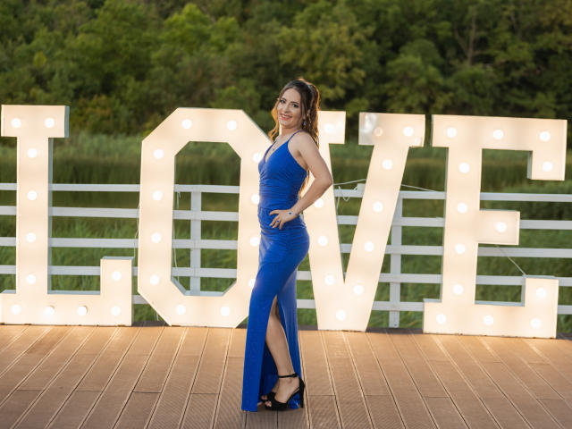 LucyLucia in a blue, ruched, sleeveless, high-slitted dress, standing on a wooden platform, wearing black heels, with a "LOVE" sign illuminated in the background.