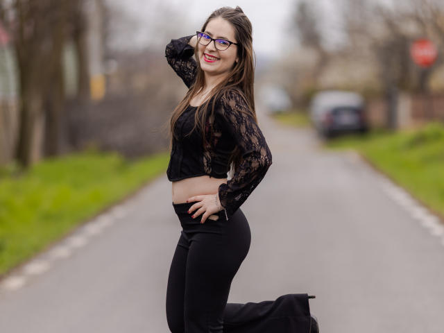 LucyLucia with long brown hair, glasses, black lace crop top, black pants, and boots, kneeling on a road, hand on head, smiling, showing her ass.