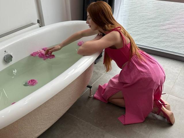 GingerFairy, with long red hair, kneels by a white bathtub, wearing a pink dress, placing pink rose petals in the water. Her ass is slightly visible.