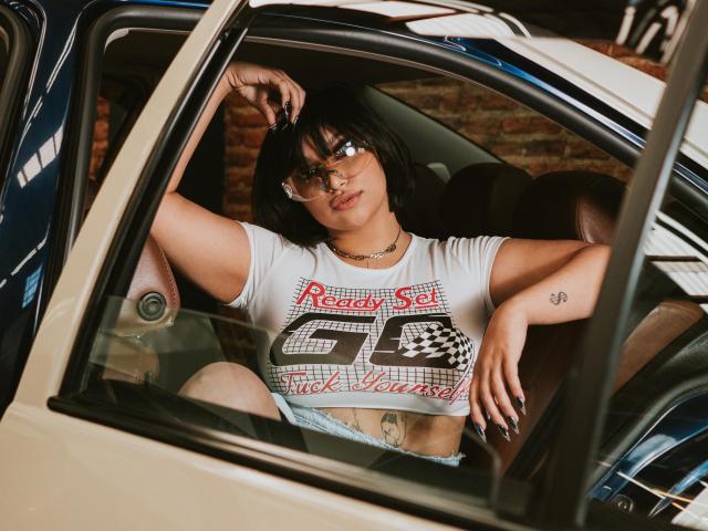 EdenSmiith com cabelo curto preto, óculos, cropped top branco, mostrando os seios, tatuagem na barriga, shorts jeans, braços apoiados na janela do carro, olhando sedutivamente para a câmera..
