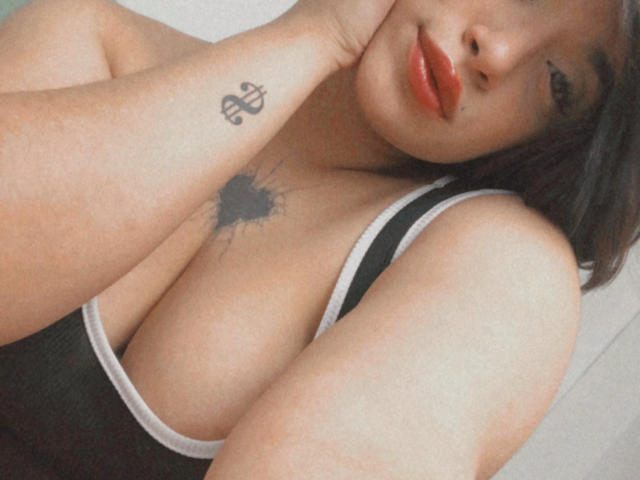 EdenSmiith with red lipstick, black and white tank top, showing her big tits, tattoo on her wrist, and black ink tattoo on her chest. She's resting her head on her hand.