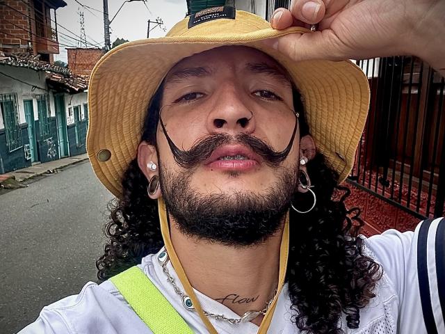 RebelDali69 with curly black hair, yellow hat, thick mustache, nose ring, earrings, hoop earring, white shirt, visible necklace, holding hat, looking up, street background.