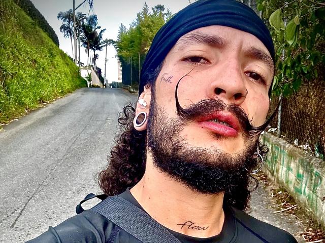 RebelDali69 with a black headband, curly beard, and mustache, wearing large ear gauges, and a black shirt, standing on a road with greenery. Tattoo on chin reads "Kill".