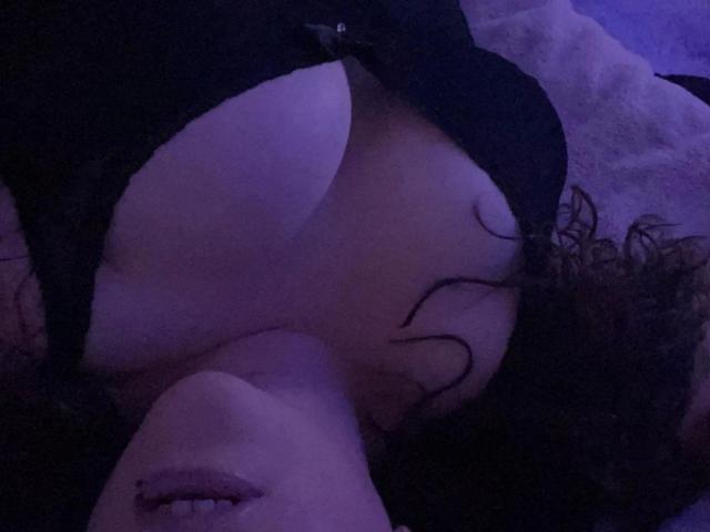 Heisse-miley-hot's boobs are visible in the top part of the image, with her face partially in the frame, showing her lips and curly brown hair. The lighting is blue.