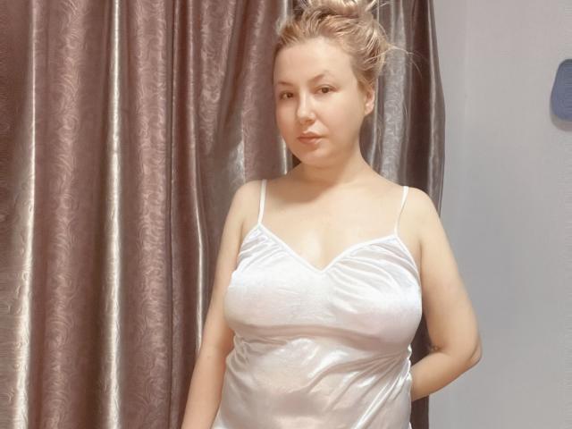HanterHellen with blonde hair in a messy bun, wearing a white, sheer, satin camisole, showing her big tits, standing in front of brown, patterned curtains.