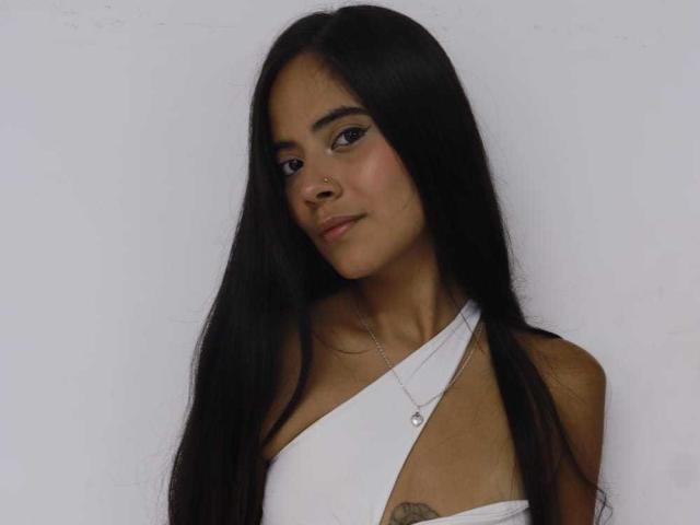 TamaraPeach has long black hair, a nose ring, and a white halter top showing a tattoo on her left boob. She's looking down with a slight smile.