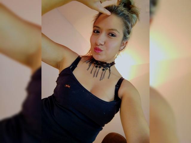 ValeriaHunter69 with blonde hair in a bun, wearing a black choker with dangling beads, black spaghetti strap dress, making a playful face, and pointing at her head.