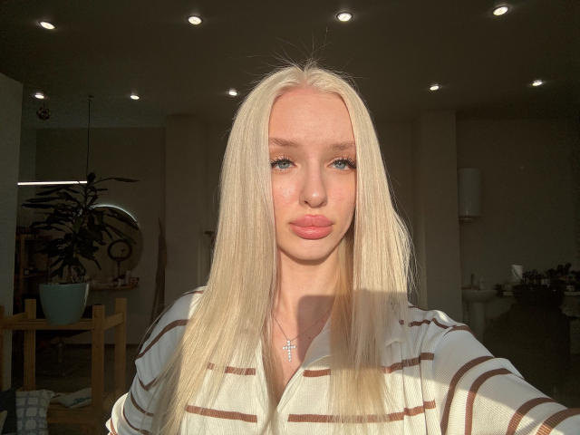 LexyGlam, met lang blond haar, blauwe ogen en roze lippen, draagt een gestreepte blouse, heeft een kruisbeeldketting om en staat in een kamer met ingebouwde plafondlampen, badend in zonlicht..